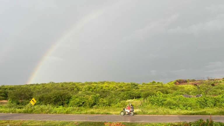 Céu nublado com arco-íris acima da estrada com uma moto passando.