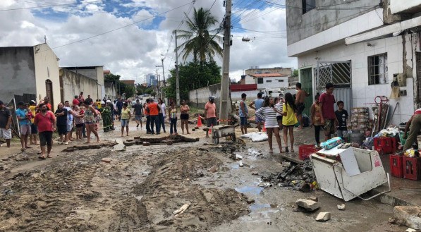 Foto tirada da cidade após desastre; dezenas de pessoas observam móveis destruídos na calçada, e rua cheia de lama.