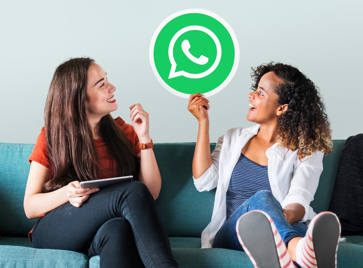 Duas mulheres conversando animadamente enquanto uma delas segura e mostra o ícone do WhatsApp.