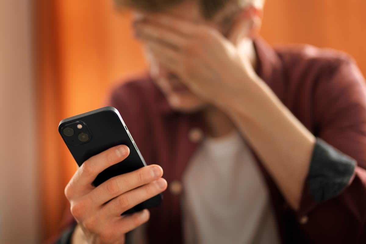 Homem segurando um smartphone na mão direita enquanto a mão esquerda está na frente do rosto em sinal de preocupação.