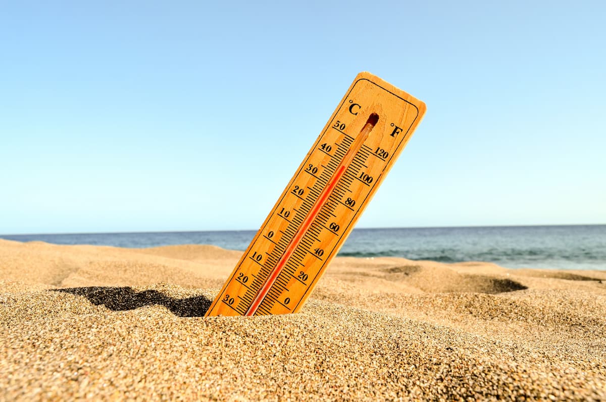 Um termômetro indicando 40ºC e 100ºF sob a areia de uma praia com céu azul e sol.
