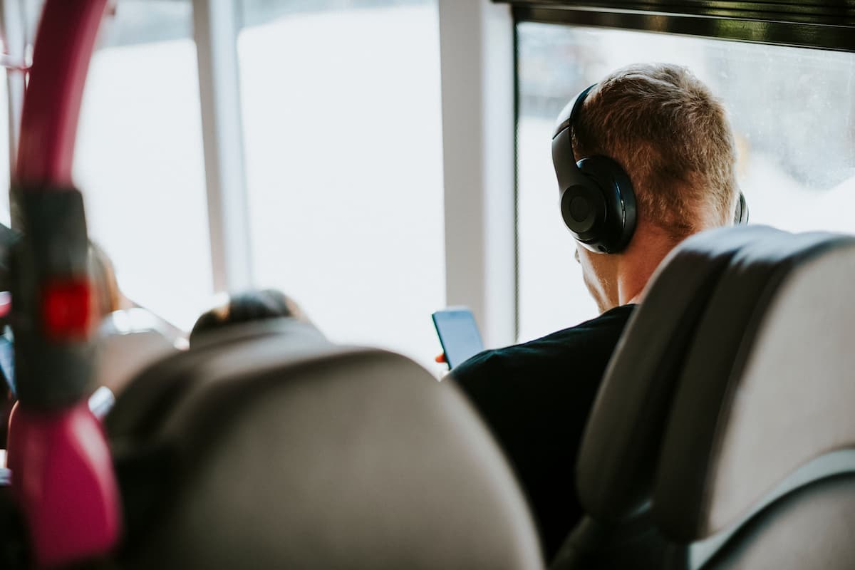 Homem sentado em poltrona de um ônibus com fones de ouvido e mexendo no seu celular.