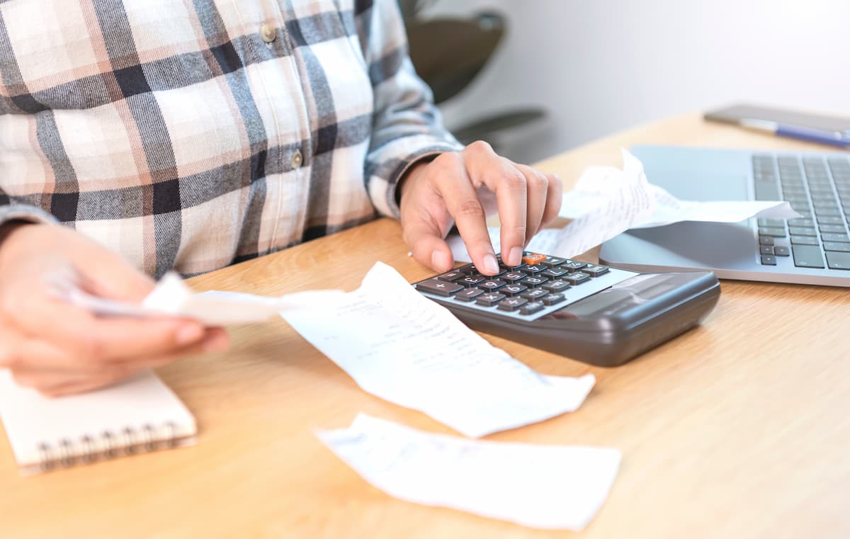 Pessoa somando contas em uma calculadora com anotações em papéis e notebook em cima de uma mesa de madeira.