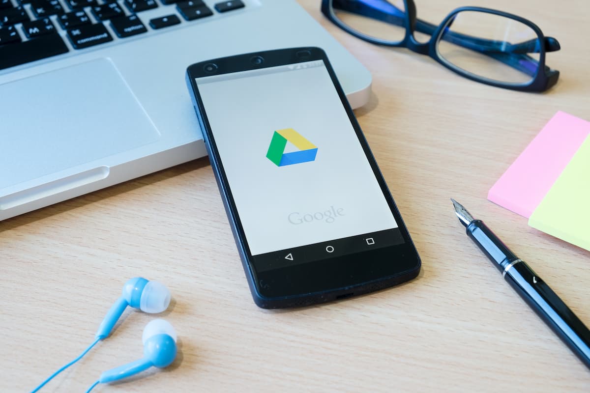 Um celular com a logo do Google Drive na tela; ele está sob uma mesa com fones de ouvidos, caneta, óculos, bloco de notas e notebook.