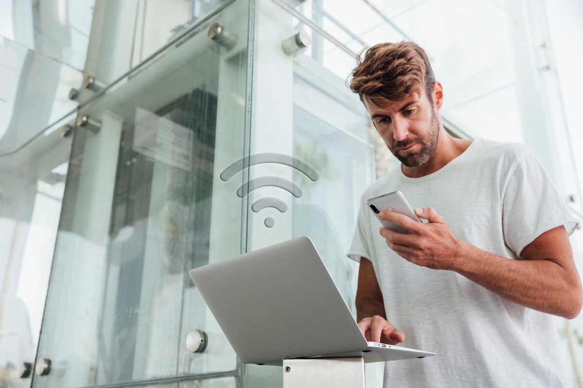 Homem usando celular e notebook, e acima dos aparelhos há o símbolo do wi-fi, adicionado digitalmente à imagem.