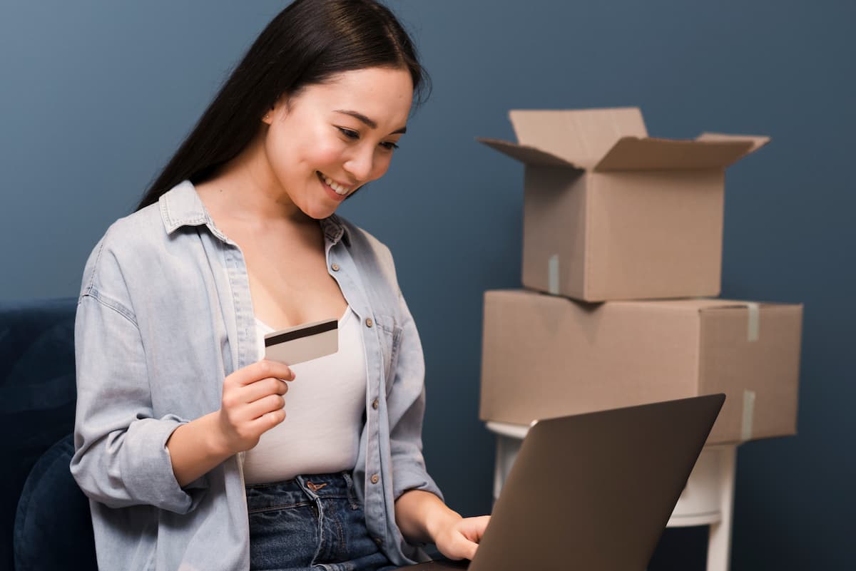 Mulher fazendo compras online com um cartão na mão direita enquanto mexe no notebook com a mão esquerda, com caixas de entregas atrás dela.