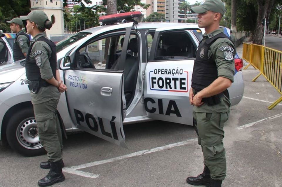Três policiais ao lado de uma viatura da Polícia de Pernambuco.
