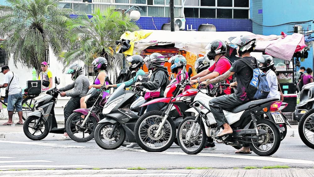 Motociclistas com capacete formam uma fileira de motos no sinal vermelho de uma rua.