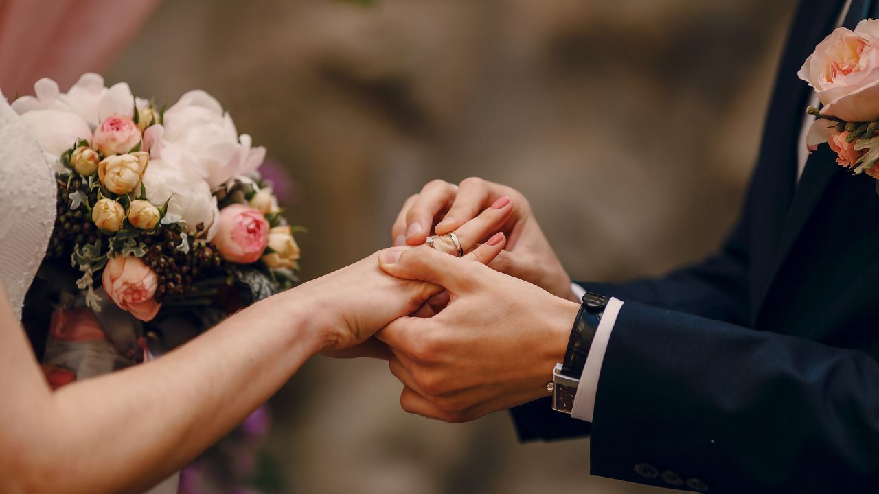 Noiva segurando um buquê de flores recebendo a aliança do noivo em seu dedo anelar.