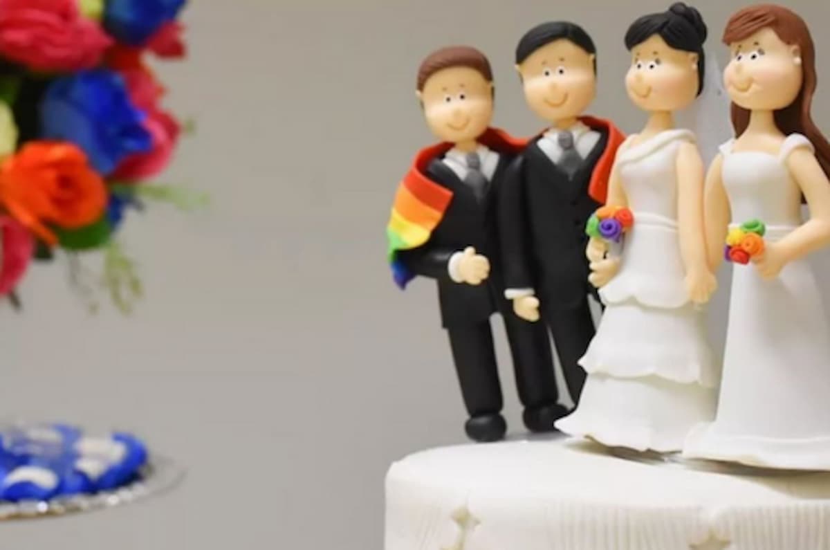 Bonecos de biscuit em cima de um bolo de casamento representando um casal homoafetivo de homens e mulheres com a bandeira do Orgulho LGBTQIA+. Ao fundo há um buquê de rosas de diversas cores.