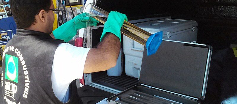 Funcionário da Agência Nacional do Petróleo (ANP) manuseando a gasolina em ensaios de vidro atrás de um carro com o porta-malas aberto.