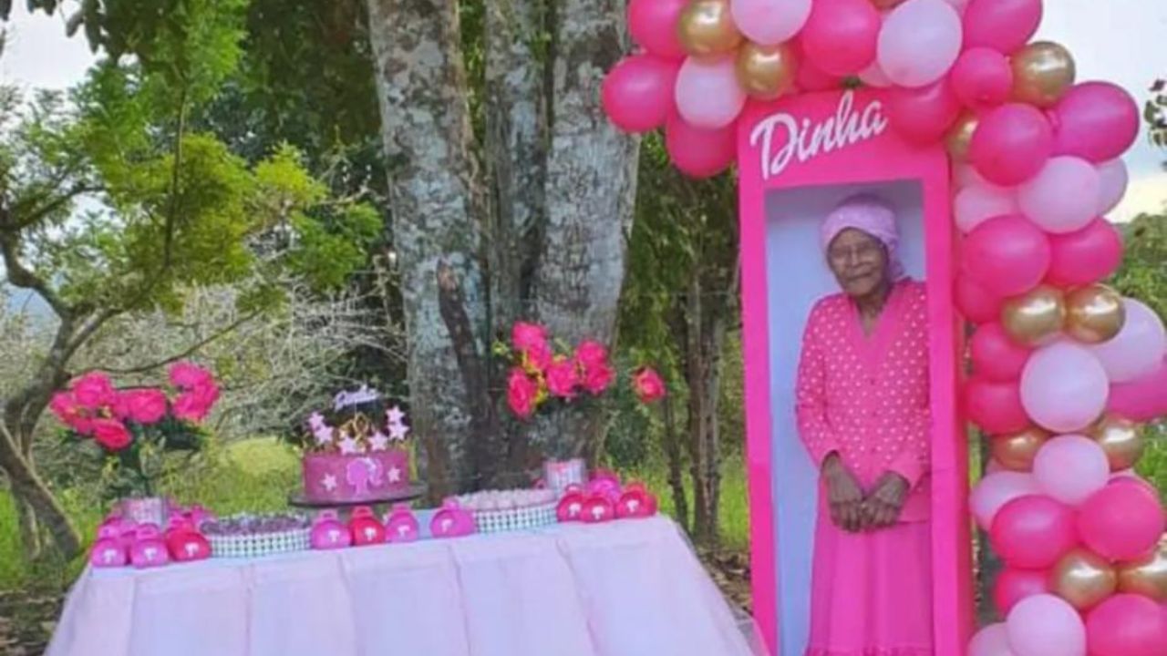 Mesa decorada com doces e bolo rosa em cima de uma toalha branca e ao lado uma caixa de boneca rosa em tamanho real com escrita em branco "Dinha", apelido de dona Josefa, que posa para a foto dentro da caixa usando um vestido e lenço rosa no cabelo.