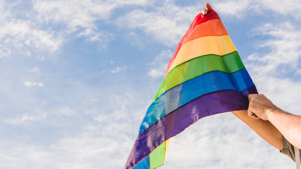 Pessoa segurando uma Bandeira Arco-íris, símbolo do movimento LGBTQIA+, no ar. Ao fundo há um céu azul com poucas nuvens.