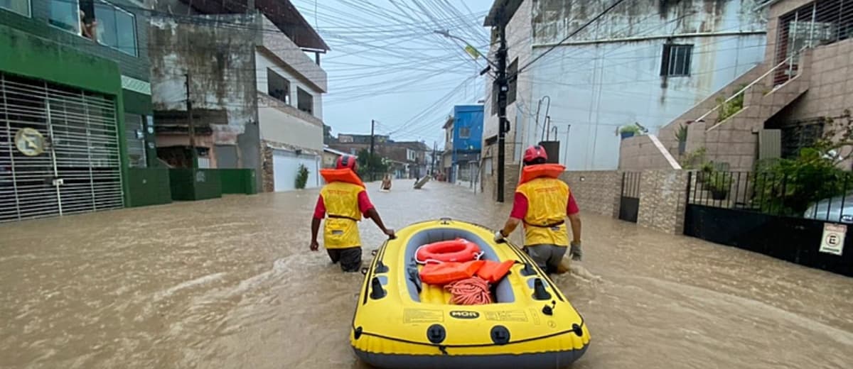 Dois agentes andando na água de uma rua inundada enquanto carregam um bote salva-vidas e outros equipamentos.