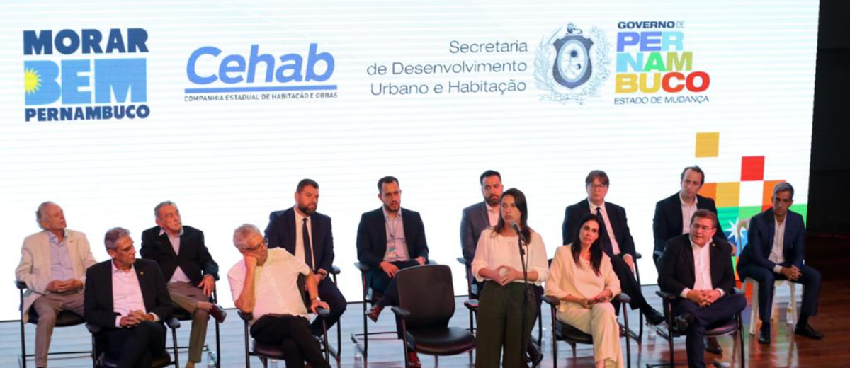 Reponsáveis pela divulgação da iniciativa de auxílio em Pernambuco para famílias de baixa renda sentados em um palco apresentando o projeto e seus patrocinadores.