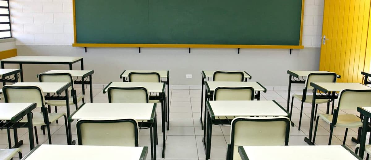 Sala de aula vazia, onde vê mesas, cadeira e lousa.