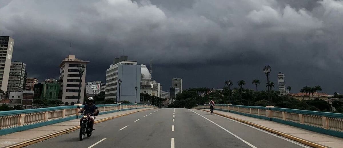 Pessoa andando de moto em avenida vazia; Ao fundo, vê-se um céu escuro, com nuvens escuras e carregadas.