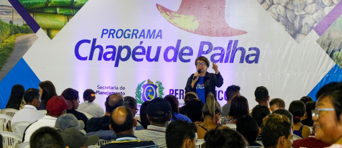 Membro do programa Chapéu de Palha, em Pernambuco, falando em microfone para diversas pessoas que estão sentadas.