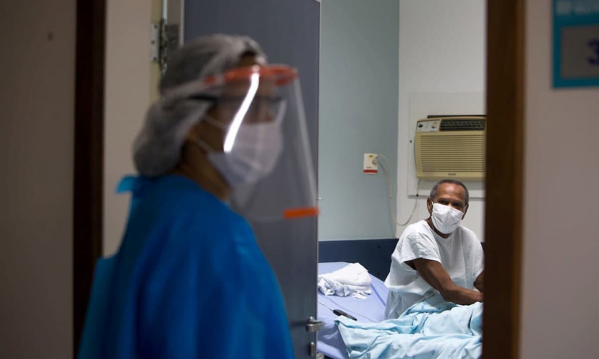 Enfermeira protegida com equipamentos de segurança médica e paciente ao fundo, sentado em uma maca no quarto de um hospital, usando uma máscara.